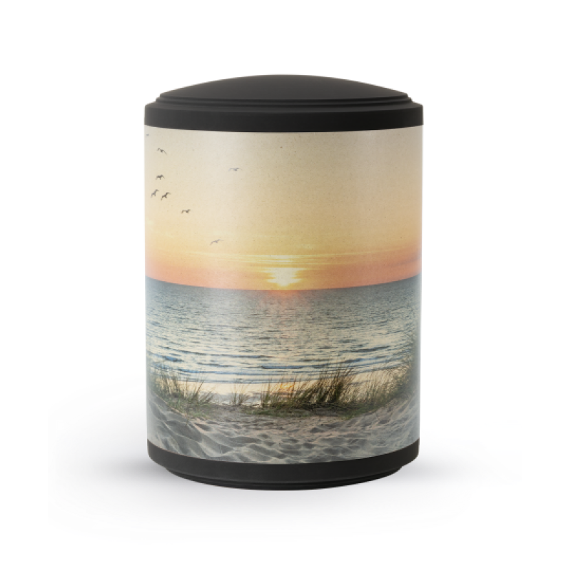 Die Urne aus der Edition Mare ist zylindrisch, schwarz mit einem farbigen, naturmotivierten Druck von Dünen und Sonnenuntergang auf der Oberfläche.
