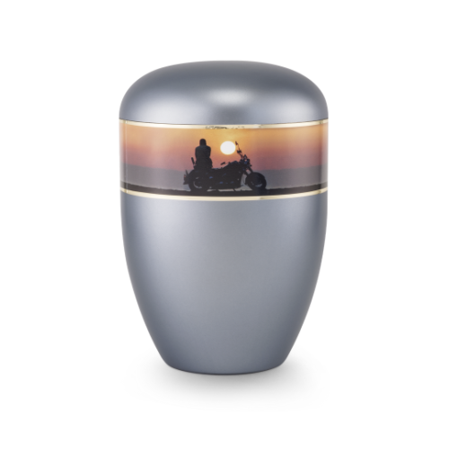 Die Urne in perlmutt stahlgrau hat eine ovale Form, einen glänzenden Deckel und eine dekorative, farbige Band mit einer Motorrad-Silhouette vor Sonnenuntergang.