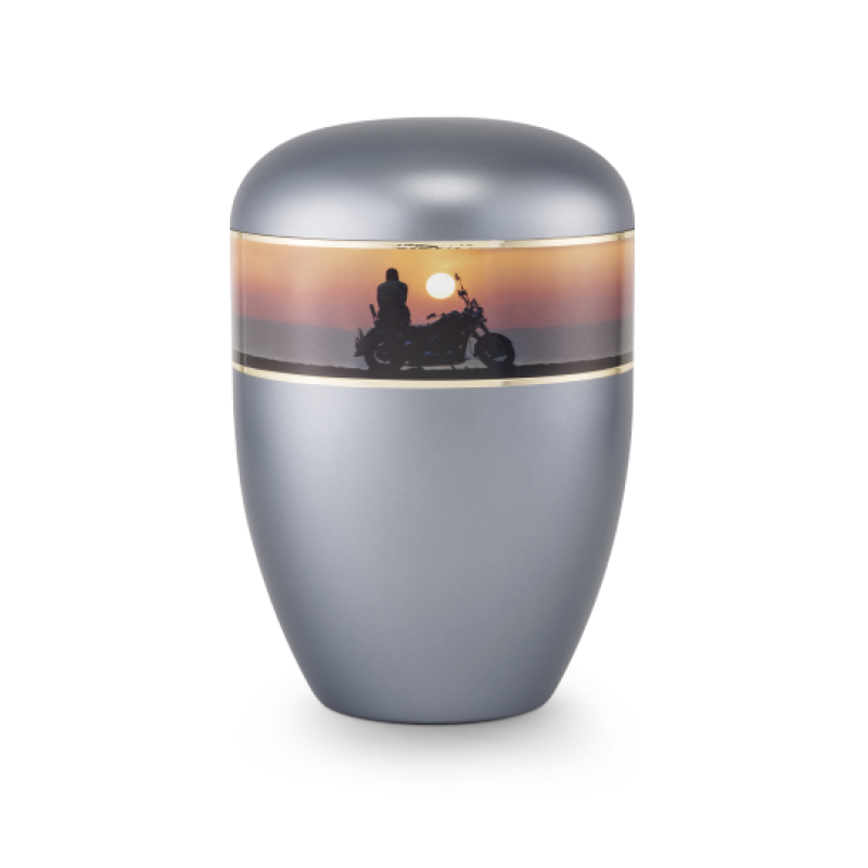 Die Urne in perlmutt stahlgrau hat eine ovale Form, einen glänzenden Deckel und eine dekorative, farbige Band mit einer Motorrad-Silhouette vor Sonnenuntergang.