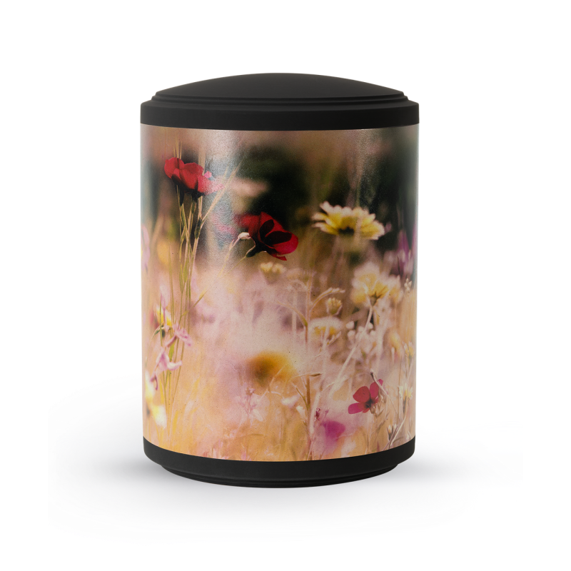 Eine schwarze Urne mit einem bunten Blumenwiesen-Motiv, aus Pflanzenfasern gefertigt, in zylindrischer Form und mit einem abnehmbaren Deckel.