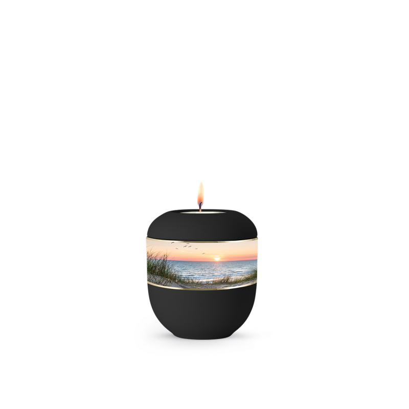 Die Urne aus der Edition Mare ist aus schwarzem Samton und zeigt eine Landschaft mit Dünen und Wasser, während oben eine Kerze brennt.