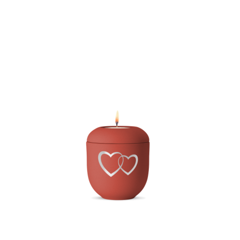 Ein rotes Gedenklicht aus Samton mit zwei ineinander verschlungenen Herzen in Weißgold, in Gesamtansicht mit brennender Kerze oben.