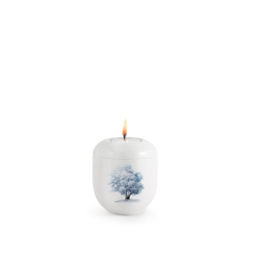 Hochglänzendes, weißes Gedenklicht mit einem blauen Winterbaum-Motiv, rundlicher Form und brennender Flamme auf der Oberseite.