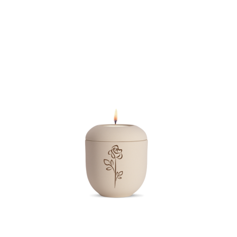 Eine cremefarbene Gedenklicht-Urne aus Keramik mit einer eingravierten Rose, Gesamtansicht mit brennender Kerze auf dem Deckel.