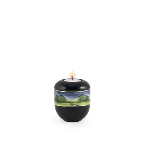 Ein schwarzes Gedenklicht in runder Form mit einem dekorativen Band, das eine Landschaft mit Flusslauf und goldenen Streifen zeigt, brennt oben.