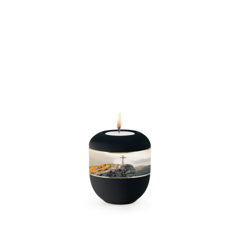 Eine schwarze Samtonurne mit umlaufendem Zierband und einem Gipfelkreuz-Motiv, aus Porzellan gefertigt, in Gesamtansicht mit brennender Kerze oben.