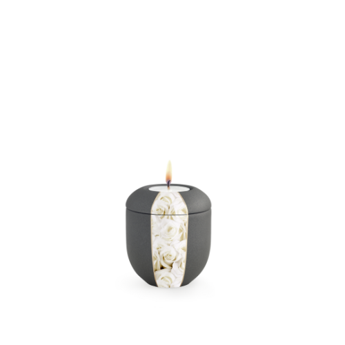 Gedenklicht in anthrazit mit Samtoberfläche, verziert mit weißen Rosen, rundliche Form, Gesamtansicht mit brennender Kerze oben.