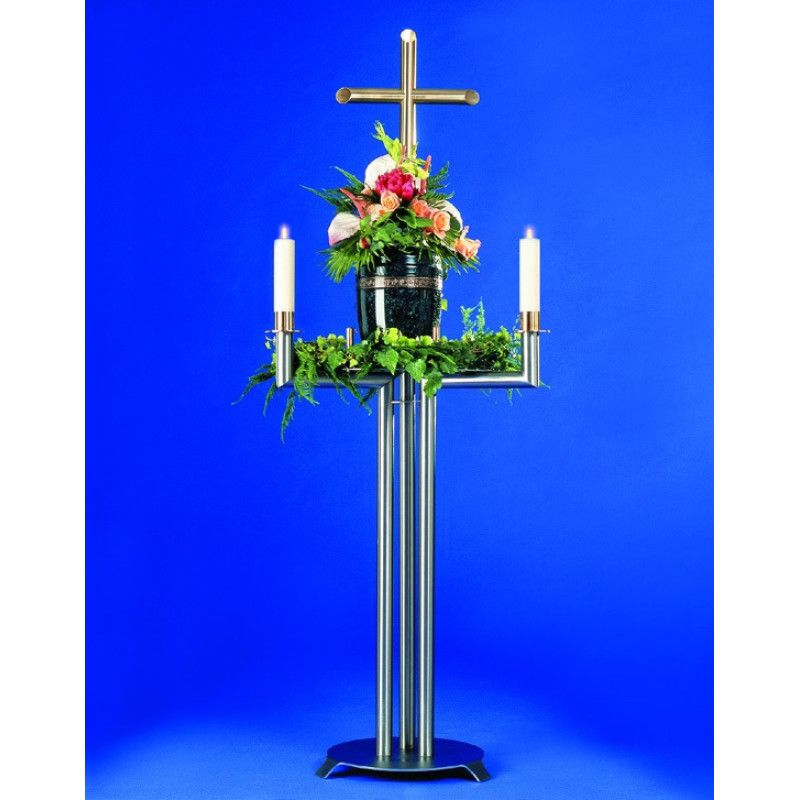 Ein Urnenkandelaber aus Edelstahl mit goldfarbener Kreuzapplikation, zwei Kerzenhaltern und einer Ablage für eine Urne, dekoriert mit Blumen und Grün.