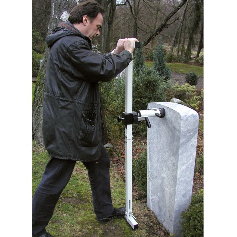 Die Handhabungshilfe ist ein silbernes, stabiles Gerät aus Metall, das zur Prüfung eines Grabsteins verwendet wird und in einer Gesamtansicht mit einer Person im Einsatz gezeigt wird.