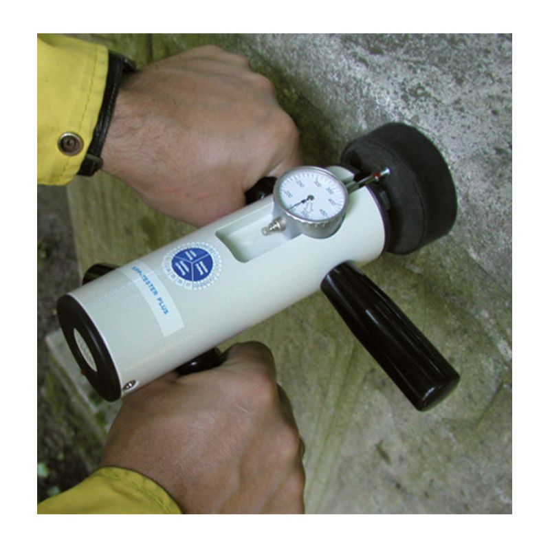 Das Bild zeigt eine Kraftmesseinrichtung in Weiß mit einem schwarzen Gummifuß, einem Manometer und einem Griff, gehalten von zwei Händen an einem Grabstein.