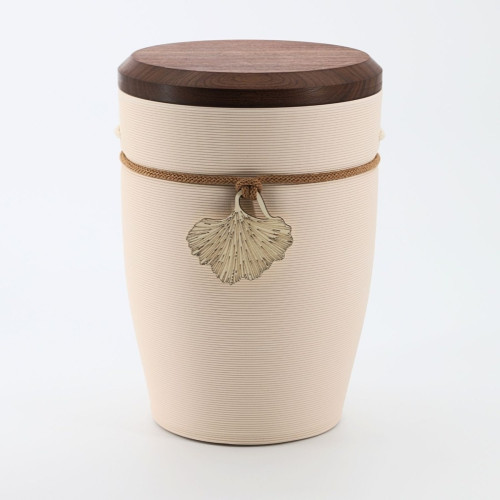 Die Urne Andenken Ginkgo in Creme ist aus Tonerde gefertigt, hat eine runde Form, eine Holzoptik auf dem Deckel und ist mit einem geflochtenen Band sowie einem Ginkgo-Anhänger verziert.