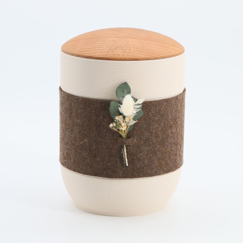 Die Urne Mira Band in Braun und Creme aus Tonerde hat eine runde Form, eine Holzdeckel und ist mit einem braunen Filzband sowie einer floralen Dekoration versehen.