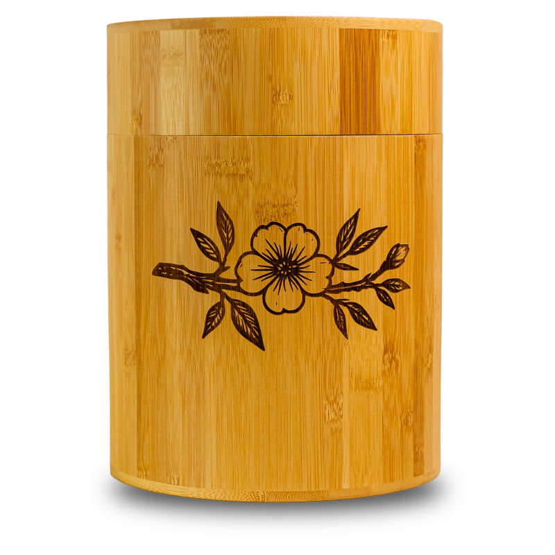 Die Bambusurne hat eine zylindrische Form, ist aus hellem Bambus gefertigt und zeigt ein eingraviertes Motiv mit einer Blume und Zweigen auf der Vorderseite.