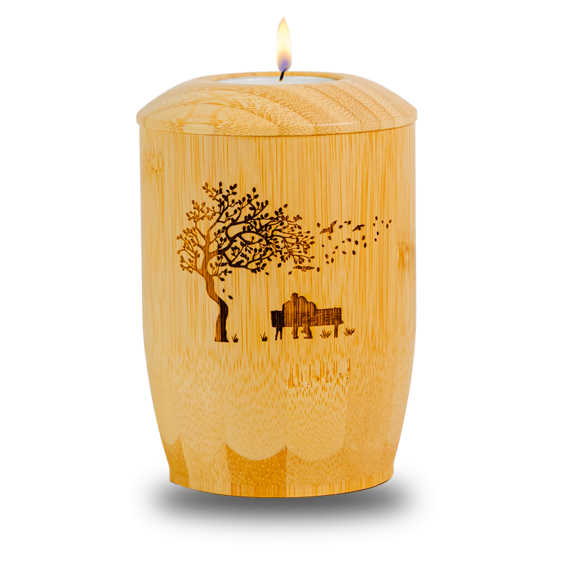 Die Bambusurne hat eine ovale Form, ist aus hellem Bambus gefertigt und zeigt ein eingebranntes Motiv mit einem Baum und einem Pärchen auf einer Bank.