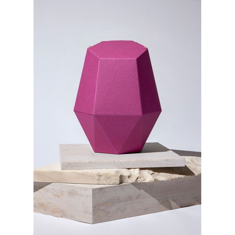 Die Kinderurne Aurora aus pinker Recycling-Kartonage hat eine geometrische Form mit einer facettierten Oberfläche und steht auf einem hellen, strukturierten Untergrund.