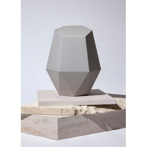 Die Kinderurne Nebel aus grauem Recycling-Kartonage hat eine facettierte, geometrische Form und steht auf einer hellen, rechteckigen Unterlage.