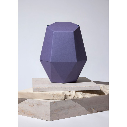 Die violette Kinderurne aus Recycling-Kartonage hat eine geometrische Form mit facettierten Flächen und steht auf einer neutralen, hellen Unterlage.