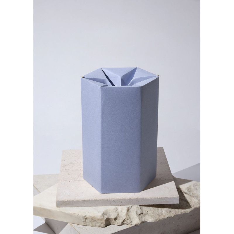 Eine hellblaue, sechseckige Papierurne aus CO2-neutralem Material mit einer gefalteten Oberseite, auf einem hellen Untergrund präsentiert.